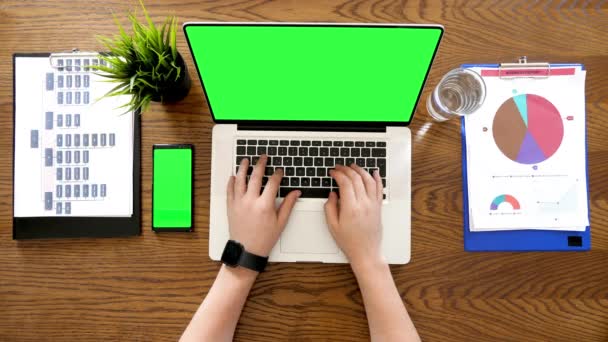 Manos masculinas escribiendo en el ordenador portátil con una pantalla verde croma al lado de un teléfono con croma en él también — Vídeos de Stock