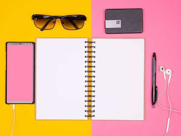 Cuaderno de papel abierto junto a un bolígrafo, gafas de sol y smartphone con auriculares sobre fondo bicolor —  Fotos de Stock