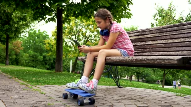 Csinos, fiatal lány, egy padon a parkban játszani vele smarphone — Stock videók