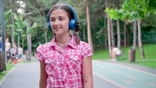 Beautiful young girl listening to music on blue headphones skates in the park — Stock Video
