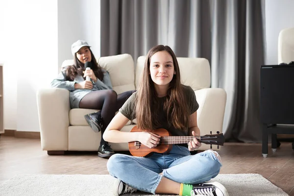 Two sisters, the younger one is playing a small guitar in front at the other is singing in the back