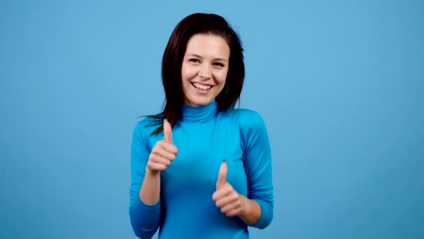 Mujer alegre alegre positiva mostrando pulgares hacia arriba sobre fondo azul — Vídeos de Stock