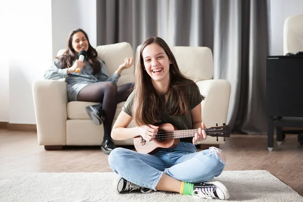Two sisters, the younger one is playing a small guitar in front at the other is singing in the back
