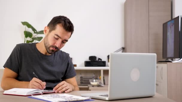 Empresário em casa em roupas casuais com um laptop na mesa toma notas em um caderno — Vídeo de Stock