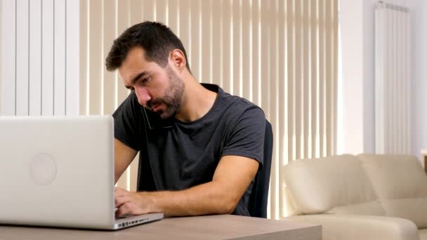 Hombre en su sala de estar hablando por teléfono y escribiendo en el ordenador — Vídeo de stock