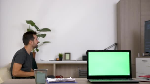 Dator med gröna mock-up skärm och en man som spelar TV-spel på konsolen suddig i bakgrunden — Stockvideo