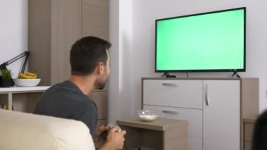 Büyük plazma Tv ekranında yeşil mock-up önünde konsolda bir video oyunu oynayan adam