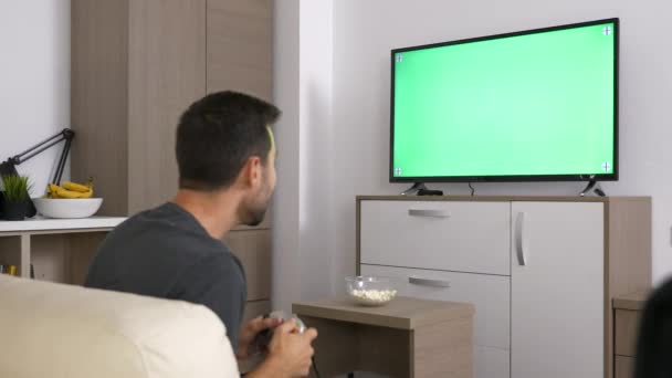 Uomo che gioca a un videogioco sulla console di fronte allo schermo mock-up verde su una grande TV al plasma — Video Stock