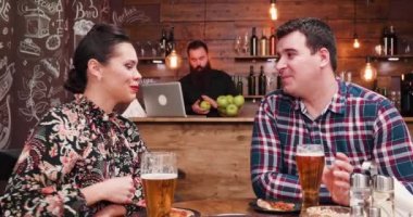 Zoom out shot of caucasian couple drinking beer and eating pizza in vintage rustic pub or restaurant