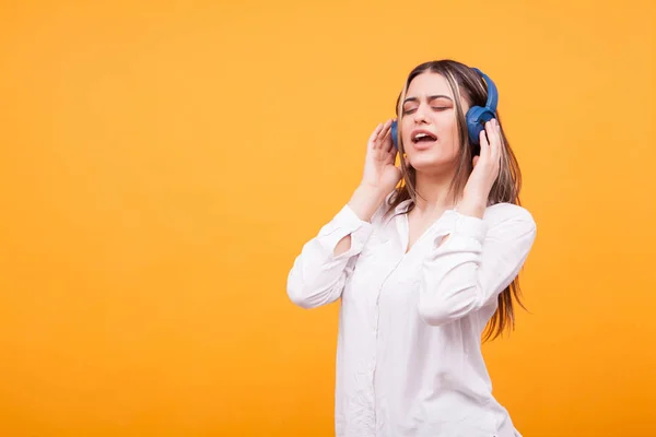 Retrato de menina animado cantando e ouvindo música em fones de ouvido azuis no fundo amarelo — Fotografia de Stock