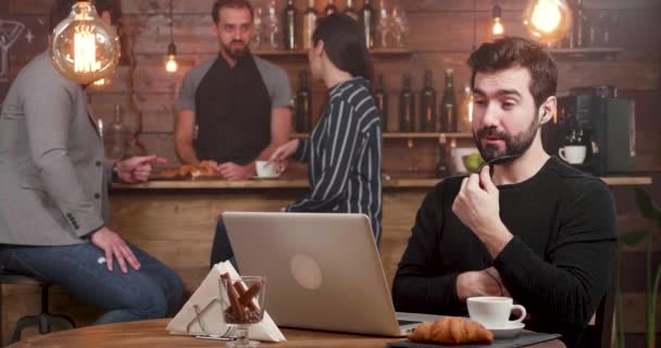 Un hombre que tiene una llamada en línea en su computadora portátil mientras trabaja en una cafetería . — Vídeos de Stock