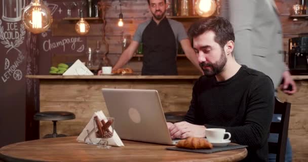Seorang pria Kaukasia dengan jenggot bekerja di laptop sementara kostumer memasuki kedai kopi — Stok Video