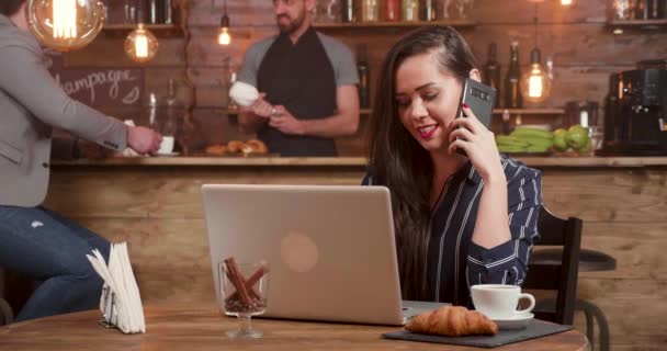 Jovencita bonita hablando en su teléfono mientras trabaja en su portátil de forma remota — Vídeo de stock