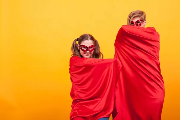 Vackra mamma med sin dotter klädd som superheros gömmer sig under deras röd cape över gul bakgrund. — Stockfoto