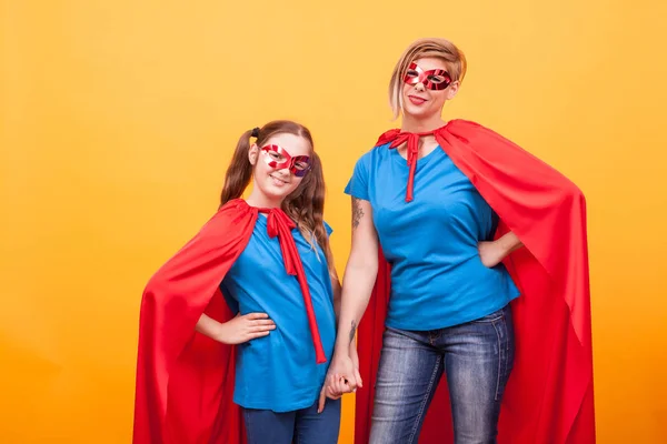 Niña y madre vestidas como superhéroes tomados de la mano y sonriendo a la cámara sobre fondo amarillo —  Fotos de Stock