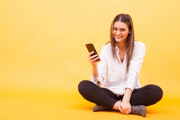 Mooi meisje haar telefoon glimlachen terwijl zitten op gele achtergrond — Stockfoto