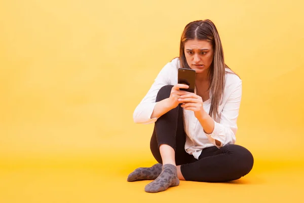 Meisje op zoek zich zorgen over haar telefoon terwijl zitten op gele achtergrond — Stockfoto