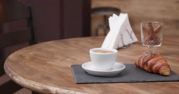 A white cup of coffee on a tray with a croissant some napkins and a glass of cinnamon — Stock Video