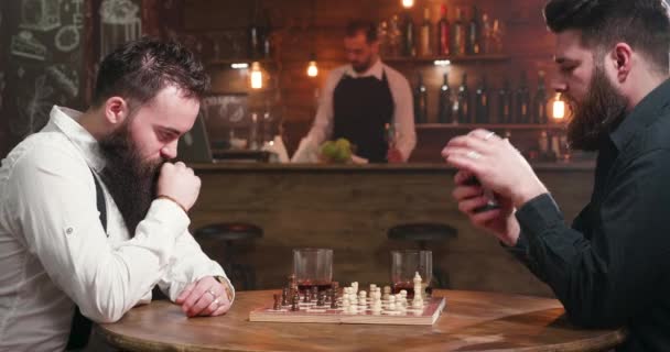 Dos amigos barbudos bebiendo y fumando mientras juegan al ajedrez en un pub — Vídeos de Stock