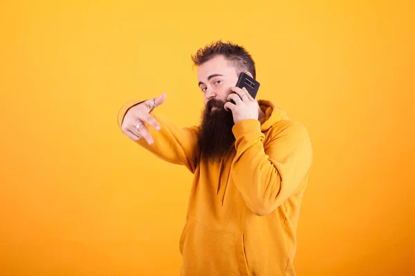 Homem na moda vestindo hoddie amarelo falando no telefone e mostrando sinal ok sobre fundo amarelo — Fotografia de Stock