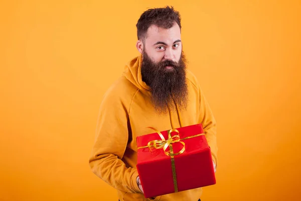 Retrato de homem engraçado com barba longa mostrando uma caixa de presente vermelha sobre fundo amarelo — Fotografia de Stock