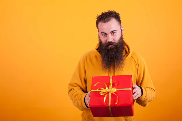 Hombre barbudo sonriente mirando a la cámara y sosteniendo una caja de regalo roja sobre fondo amarillo . —  Fotos de Stock