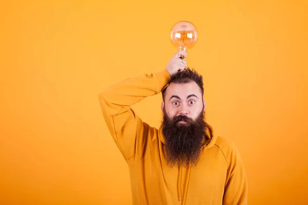 Hombre barbudo inteligente sosteniendo la bombilla en su cabeza sobre el fondo amarillo —  Fotos de Stock