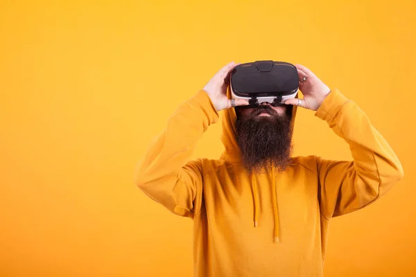 Hispter with long beard wearing yellow hoodie and virtual reality headset over yellow background