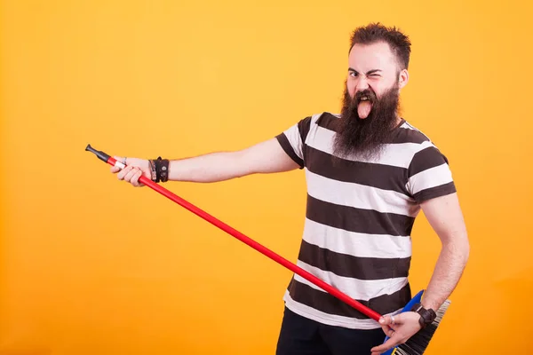Handsome bearded young man picking up a broom as if it was a guitar over yellow background. — Stock Photo, Image