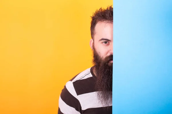 Bonito hipster barbudo escondido atrás de um painel azul sobre fundo amarelo — Fotografia de Stock