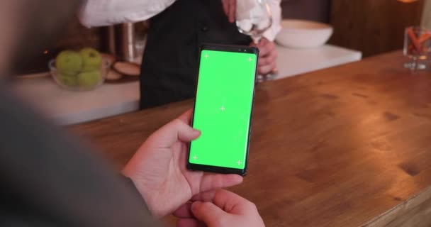 Человек ворует по телефону с зеленым экраном, сидя за барной стойкой — стоковое видео