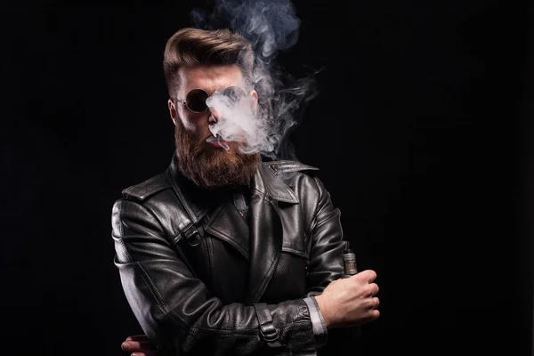 Homem barbudo elegante em couro jack fumar cigarro eletrônico sobre fundo preto — Fotografia de Stock