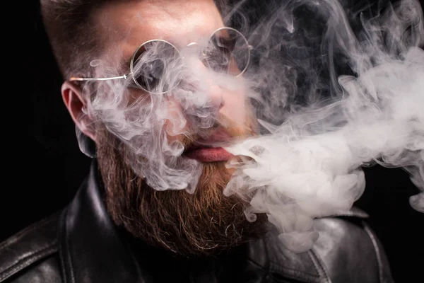 Gros plan portrait de jeune homme barbu avec des lunettes de soleil expirant fumée de cigarette sur fond — Photo