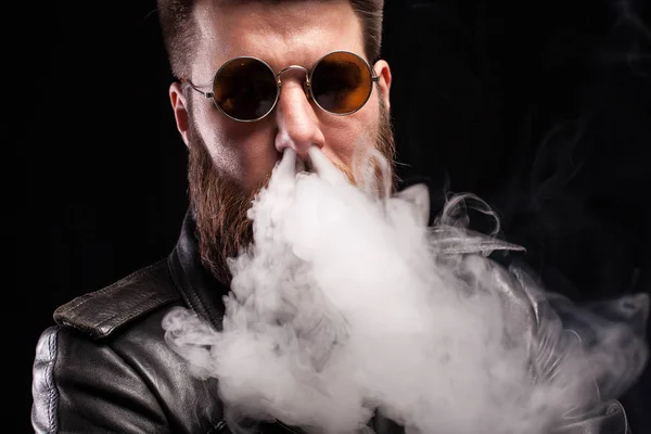 Attraente uomo barbuto espirando fumo di sigaretta sul naso su sfondo nero — Foto Stock