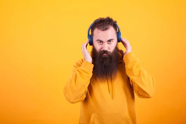 Bonito homem barbudo olhando para a câmera e ouvindo música em fones de ouvido azuis sobre fundo amarelo — Fotografia de Stock