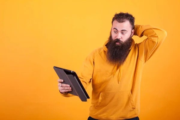 Hombre joven guapo con barba larga mirando confundido en la tableta sobre fondo amarillo —  Fotos de Stock