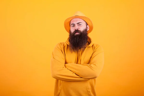 Homem barbudo com chapéu laranja em seu chapéu segurando seus braços cruzados sobre fundo amarelo — Fotografia de Stock