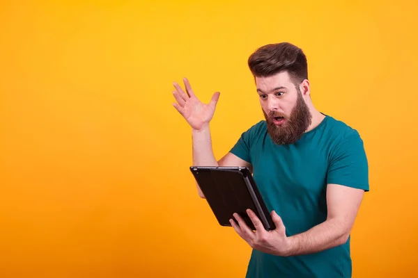 Schöner bärtiger Mann in grünem T-Shirt, der schockiert auf Tablet vor gelbem Hintergrund schaut — Stockfoto