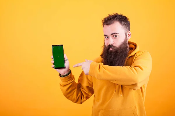 Hombre hipster barbudo intrigado apuntando a teléfono inteligente sobre fondo amarillo —  Fotos de Stock