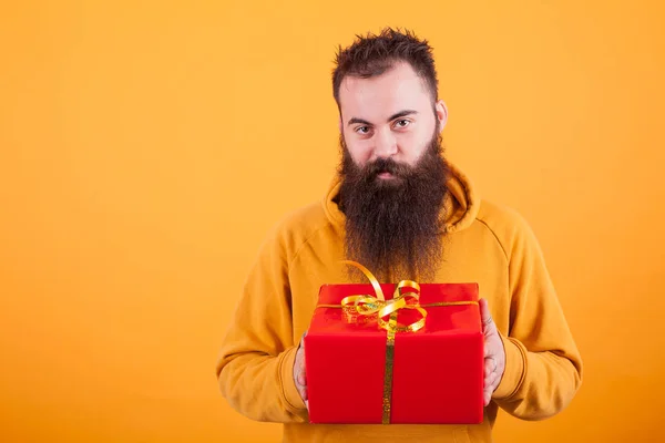 Jovem alegre com barba longa segurando uma caixa de presente vermelha sobre fundo amarelo — Fotografia de Stock