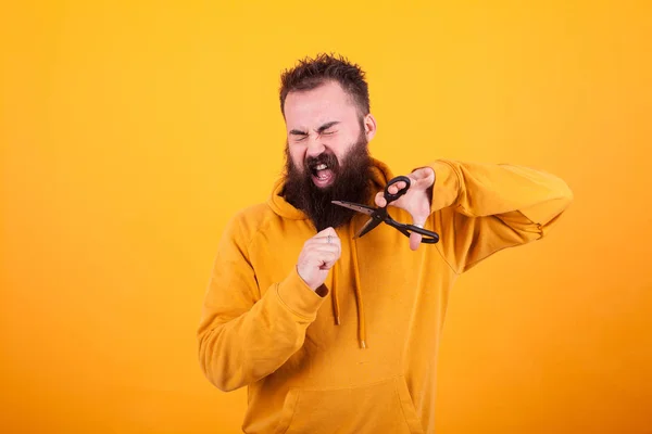 Schöner bärtiger Mann schließt die Augen, während er seinen Bart mit einer Schere über gelbem Hintergrund schneidet — Stockfoto