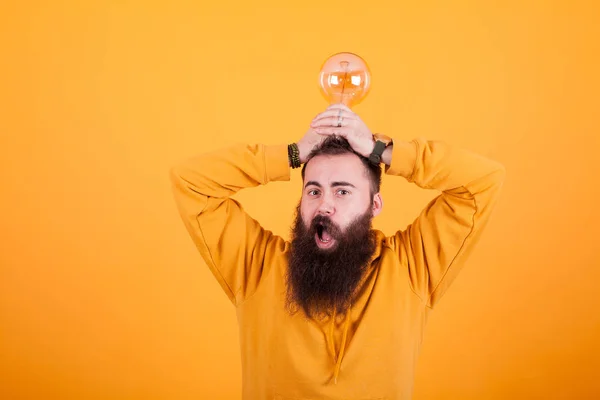 Homem barbudo olhando espantado com lâmpada sobre a cabeça na frente do fundo amarelo — Fotografia de Stock