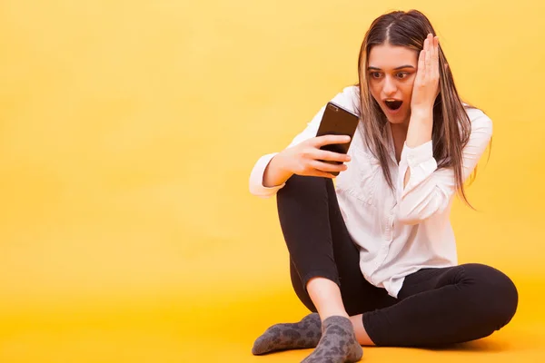 Flickan tittar förvånad över hennes telefon när du sitter över gul bakgrund — Stockfoto