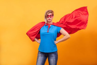 Superheros genç kadın kılık ayakta gururla kırmızı pelerinini hava ile sarı arka plan üzerinde