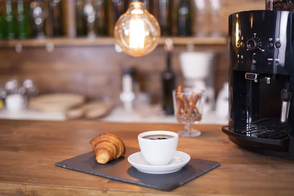 Café servi avec croissant à côté de la machine à café noire sur le comptoir du bar — Photo