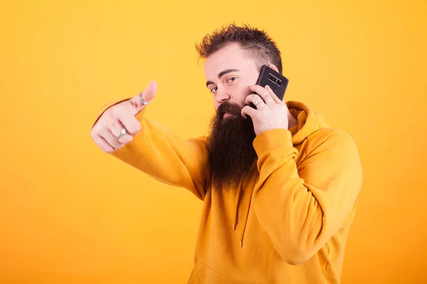 Jeune homme attrayant avec une longue barbe parlant au téléphone et montrant pouce vers le haut signe sur fond jaune — Photo