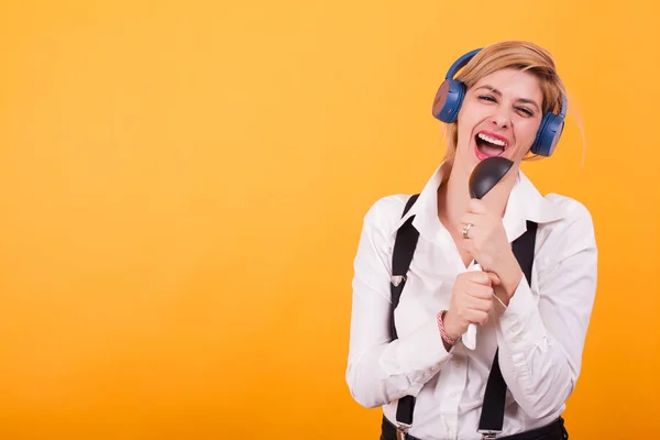 Aantrekkelijke jonge vrouw zingen op een zwarte lepel met koptelefoon op haar hoofd op gele achtergrond. — Stockfoto