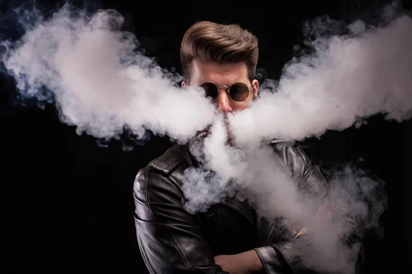 Fashionable bearded man with stylish glasses in leather jacket vaping electronic smoke.