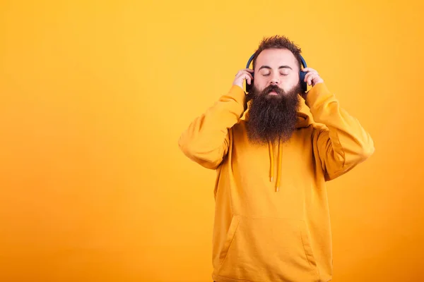 Jeune homme barbu portant un sweat à capuche jaune, écoutant de la musique sur son casque bleu paisiblement sur fond jaune — Photo