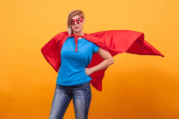 Mujer joven en traje de superhéroes de pie orgullosamente con su capa roja en el aire sobre fondo amarillo —  Fotos de Stock